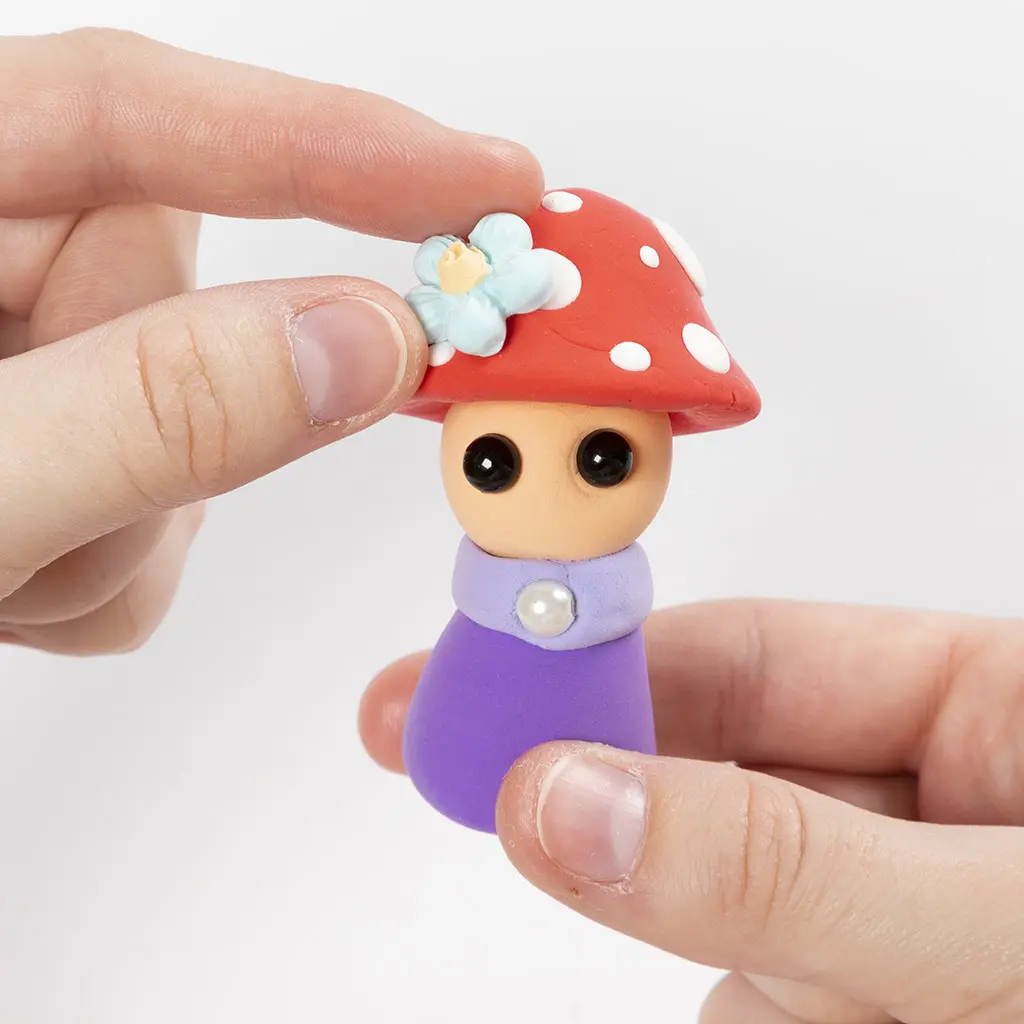Een hand plaatst een lichtblauw kleibloemetje als afwerking op de rode hoed van het paddenstoelfiguurtje.