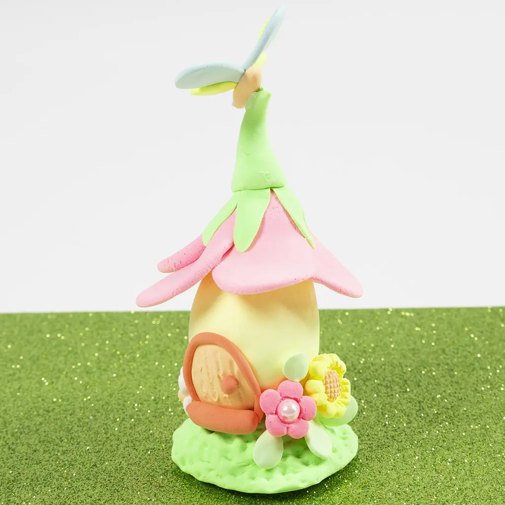 Voltooid pastelkleurig sprookjeshuisje van klei met een dak van roze bloemblaadjes, een libelle als piek, een klein houten deurtje en minibloemen, rustend op een getextureerde groene basis.