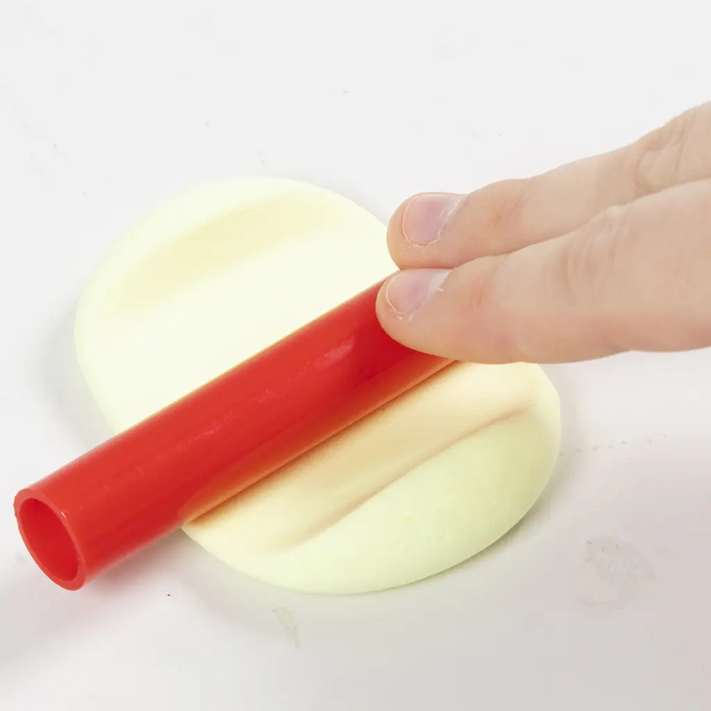 Het platrollen van een lap lichtgele klei met een rode plastic roller op een wit werkoppervlak.