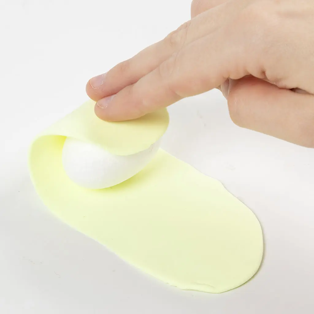 Het wikkelen van de platgedrukte lap lichtgele klei om een witte piepschuim eivorm.