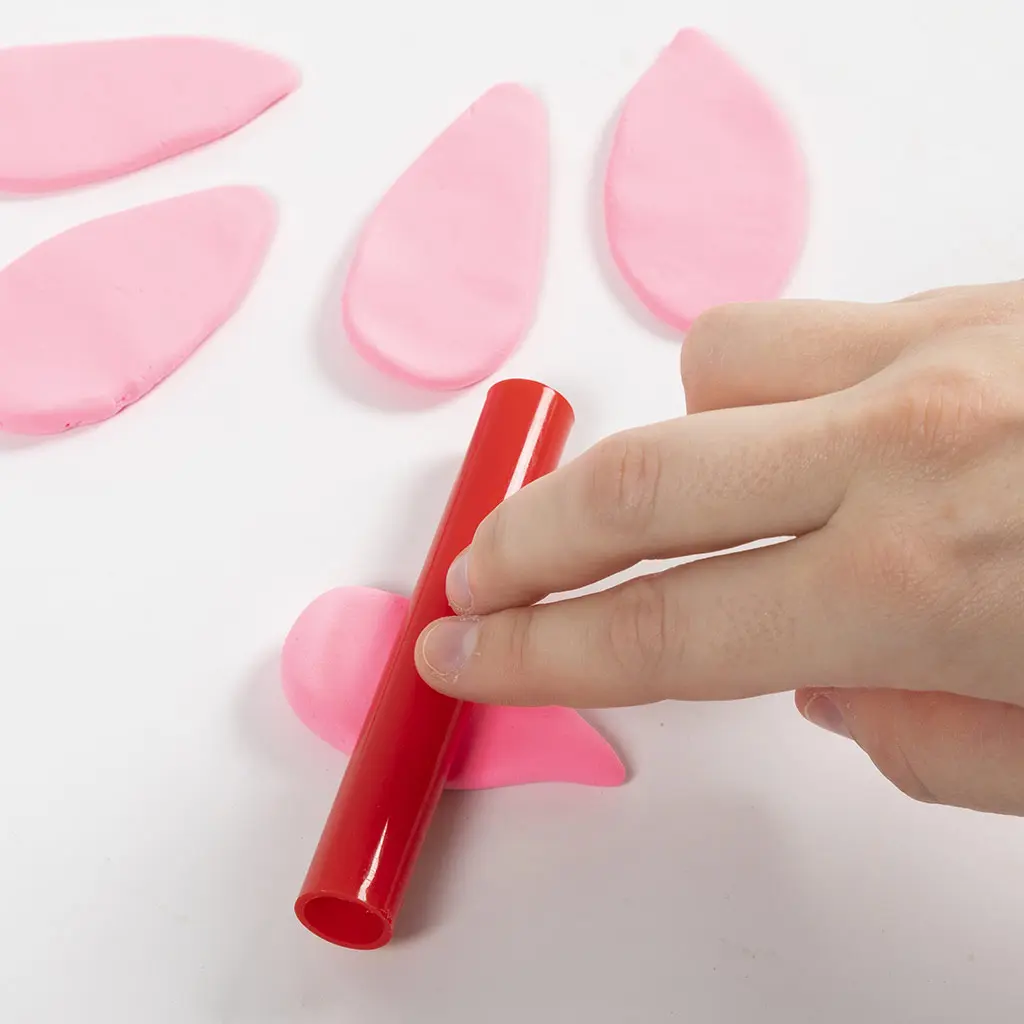 Een hand rolt roze klei plat met een rode plastic roller om bladeren voor het dak van het sprookjeshuisje te maken, met meerdere al gevormde roze kleiblaadjes op de achtergrond.
