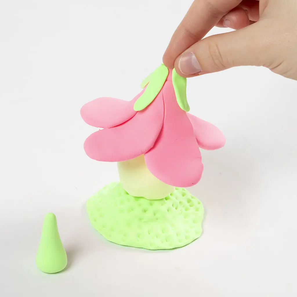 Een hand bevestigt de kleine lichtgroene kleiblaadjes over de top van het roze bloemendak op het huisje.