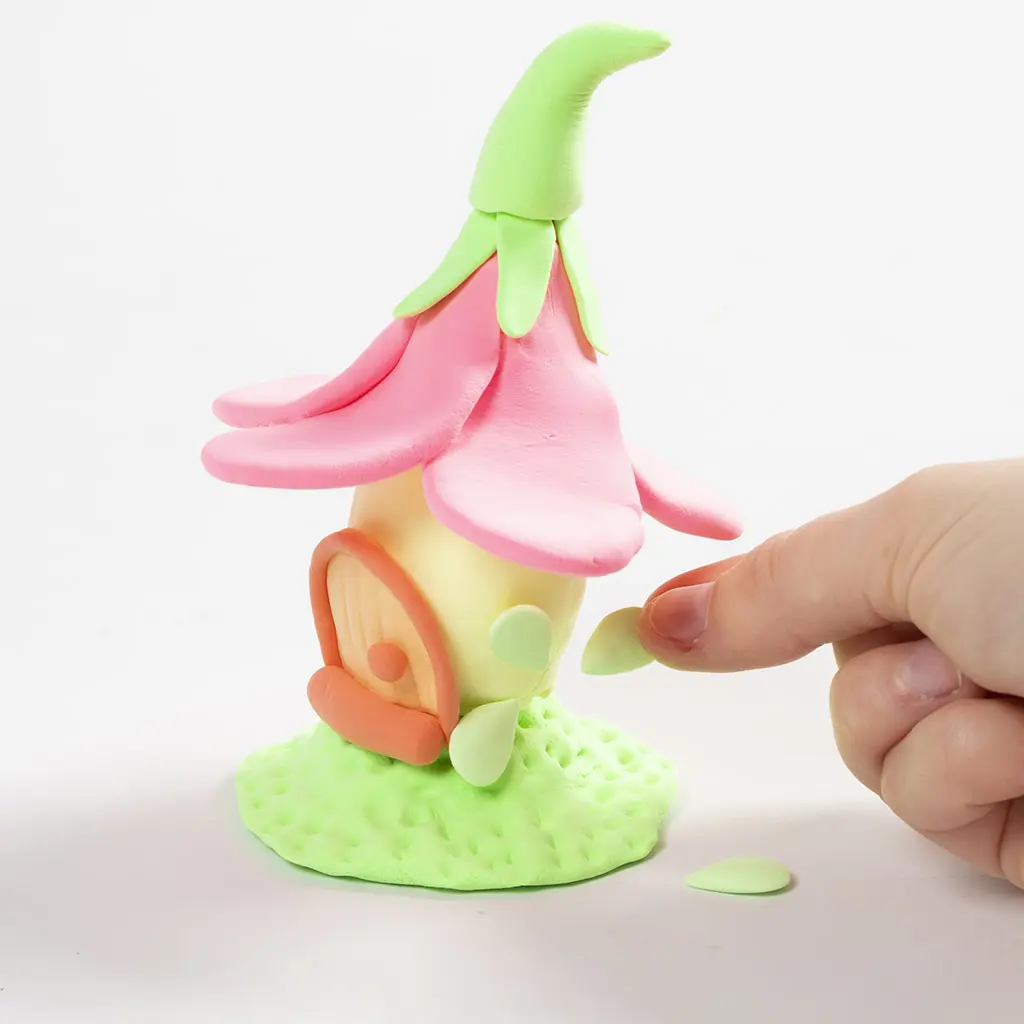 Een hand plaatst kleine groene kleiblaadjes op het gele huisje rondom het deurtje.