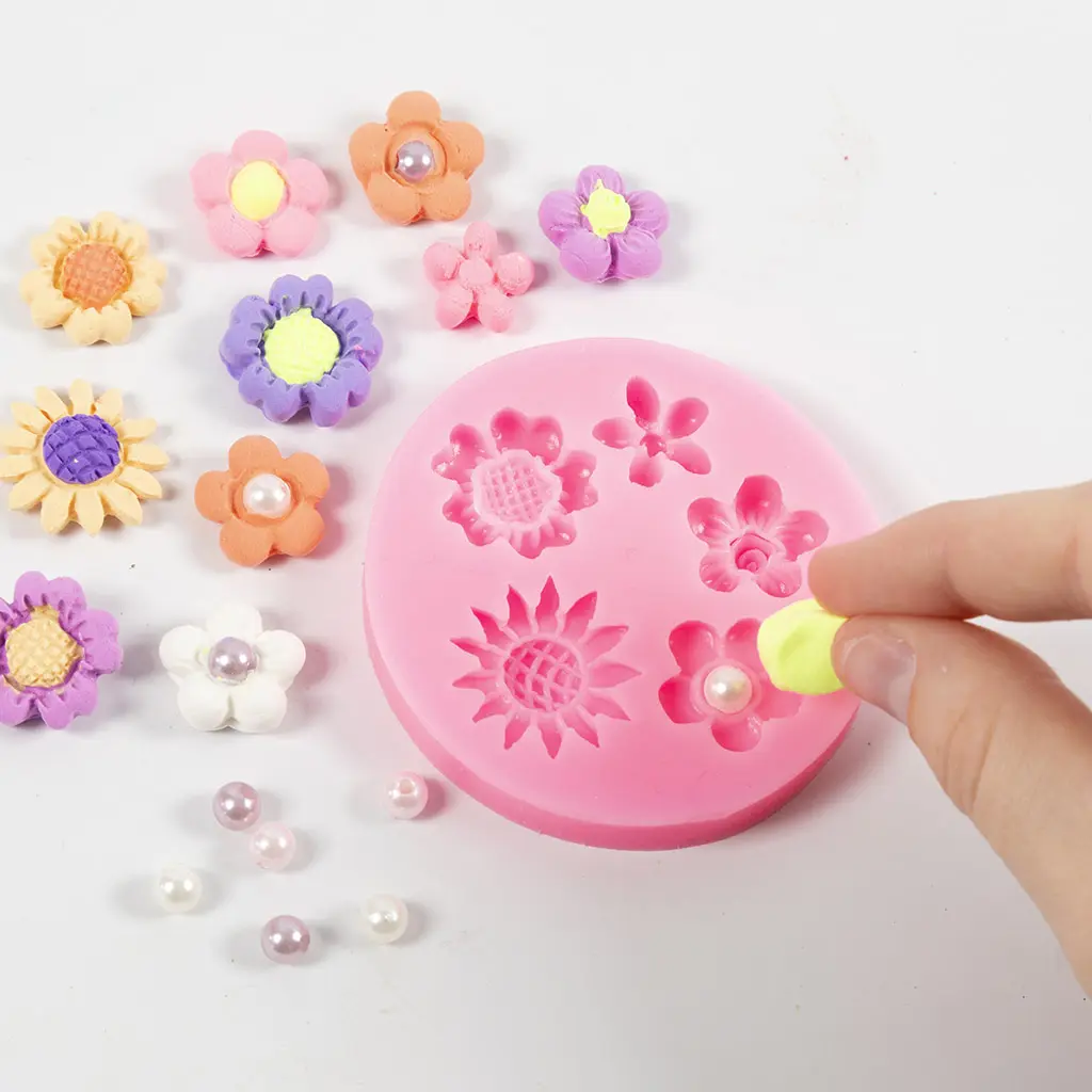 Een hand duwt gele klei in een roze siliconen gietvorm voor bloemen, met eromheen verschillende gekleurde kleibloemetjes en pareltjes.