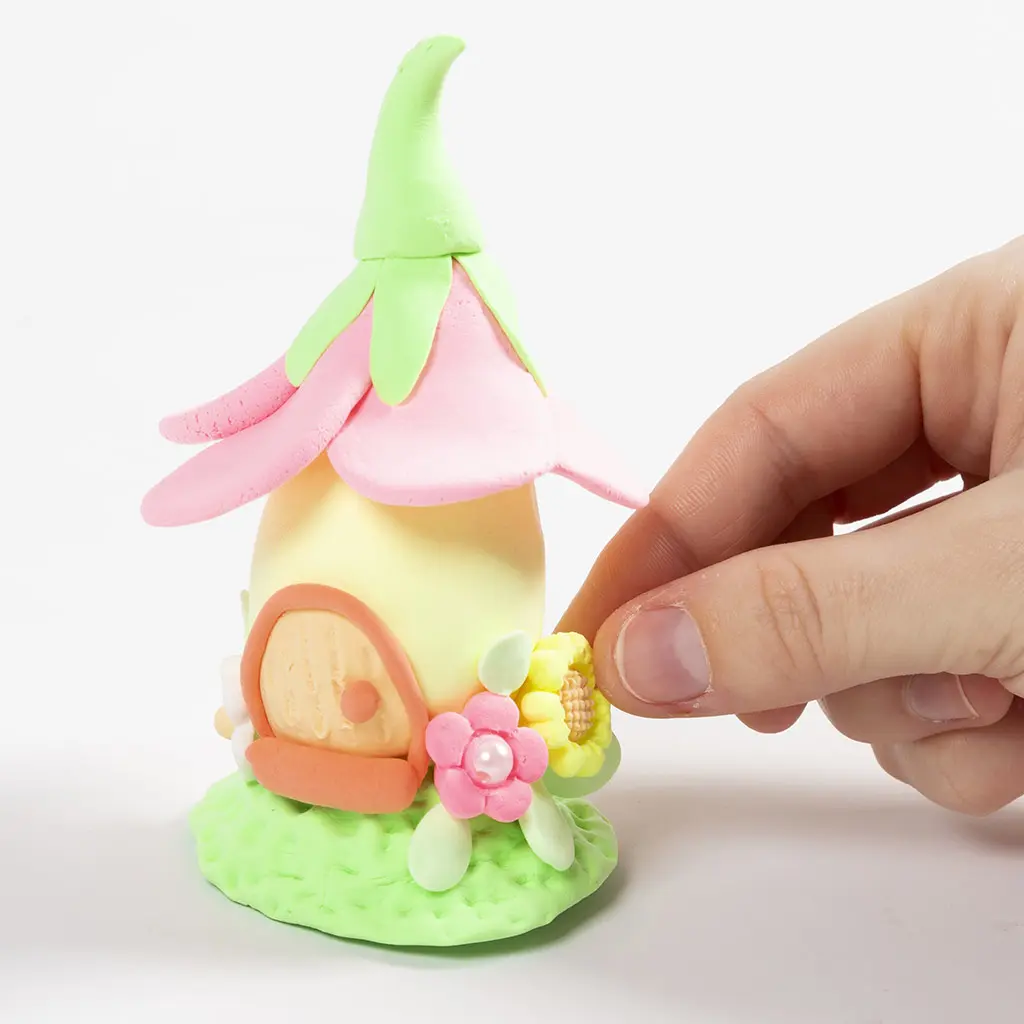 Een hand bevestigt een klein geel kleibloemetje aan de zijkant van het sprookjeshuisje bij de voordeur.