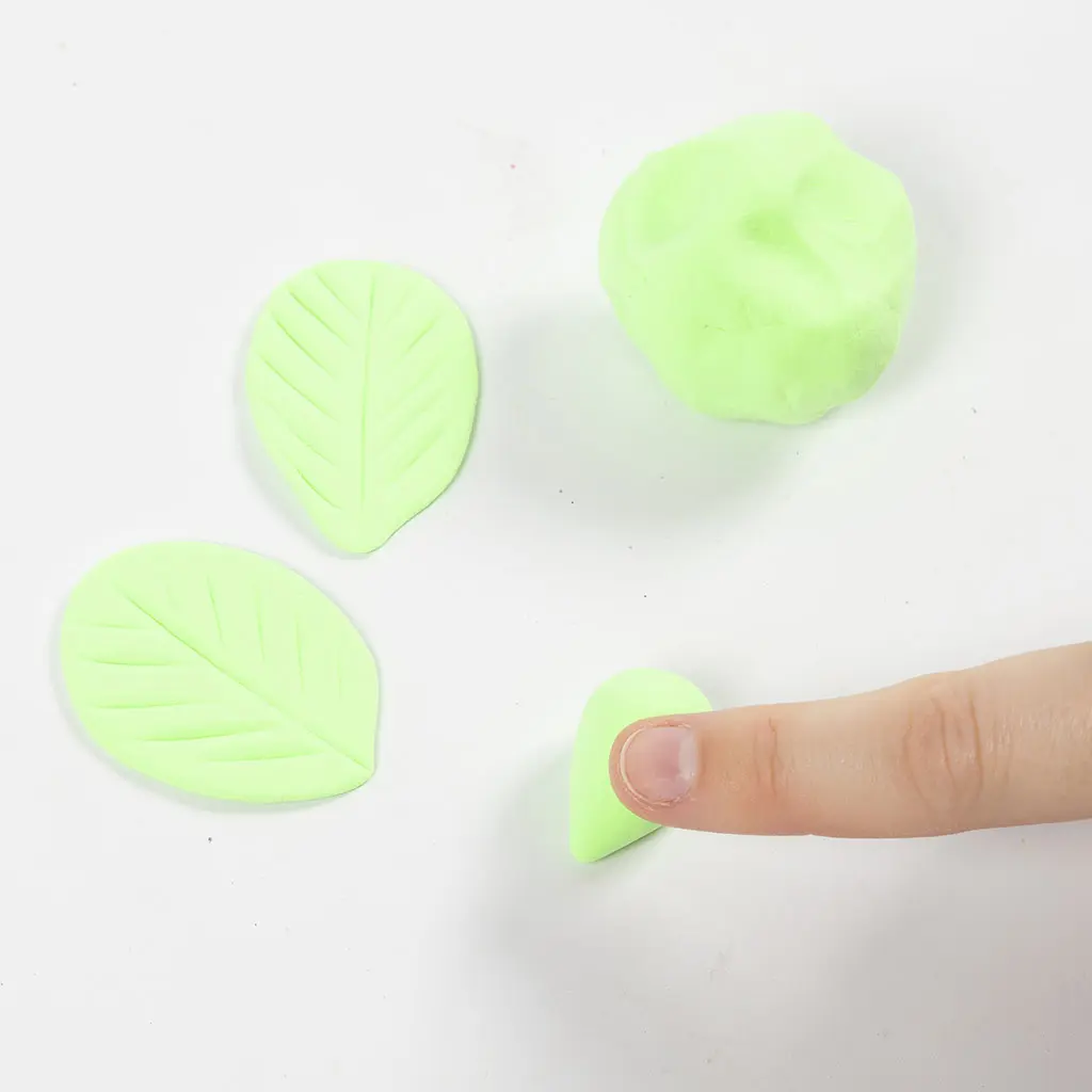 Een vinger vormt een bolletje lichtgroene klei tot een druppelvorm voor een blad van het aardbeienhuisje, met twee al afgewerkte bladeren ernaast.