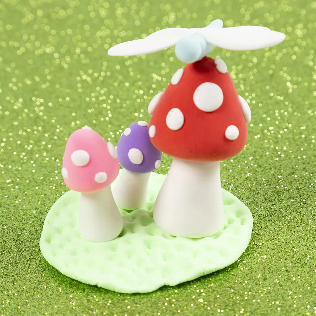 Drie kleipaddenstoelen (roze, paars en rood) met witte stippen op een getextureerde groene kleibasis, gekroond met een kleine libel op de grootste paddenstoel.
