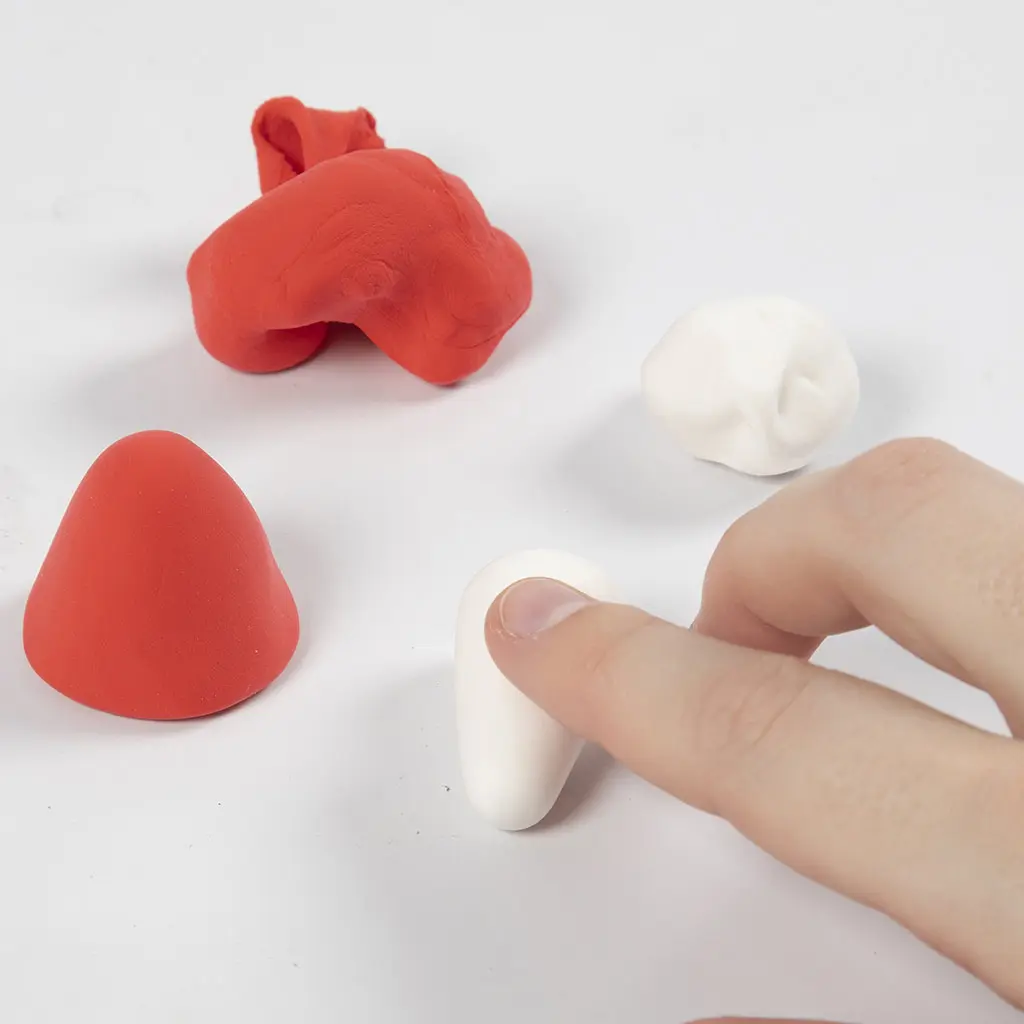 Een vinger vormt een kegeltje van witte klei voor het steelpootje van de paddenstoel, naast een rode kleihoed en losse klei.