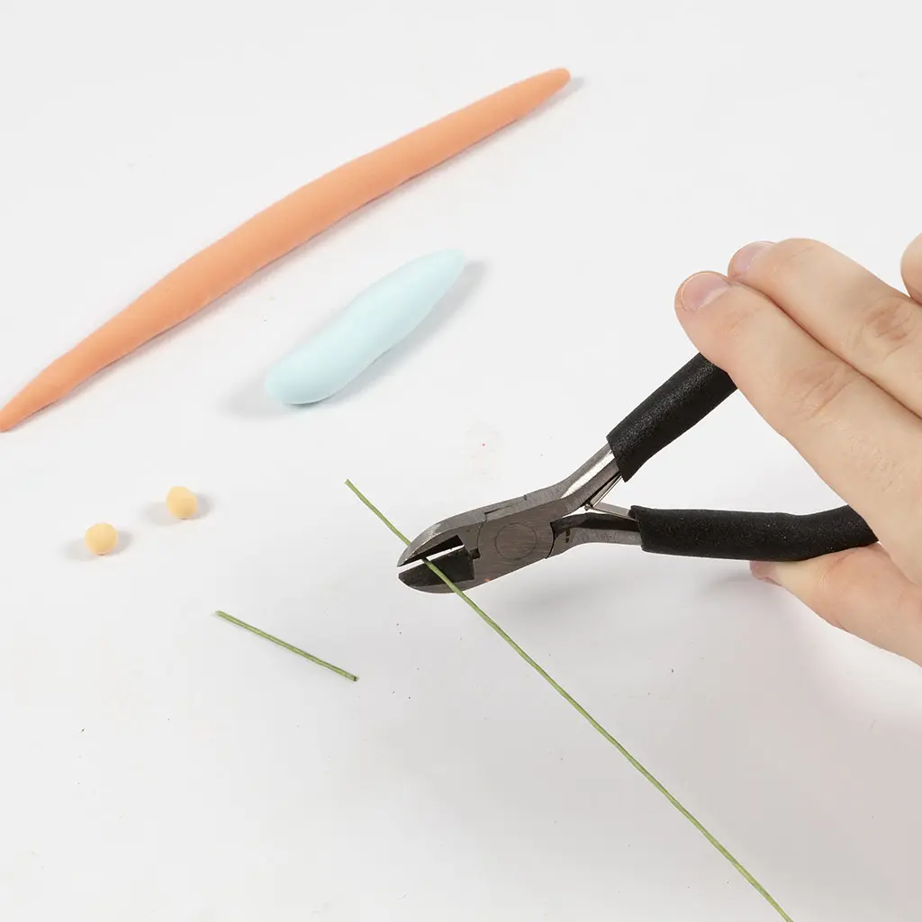 Een hand knipt een stukje dun groen wikkeldraad af met een kniptang, met op de achtergrond een uitgerolde oranje kleislang, een blauw kleilijfje en twee gele bolletjes.