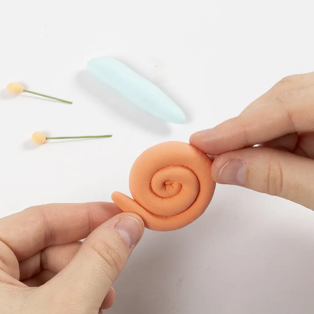 Twee handen rollen een lange oranje kleislang op tot een spiraal om het slakkenhuis te vormen.