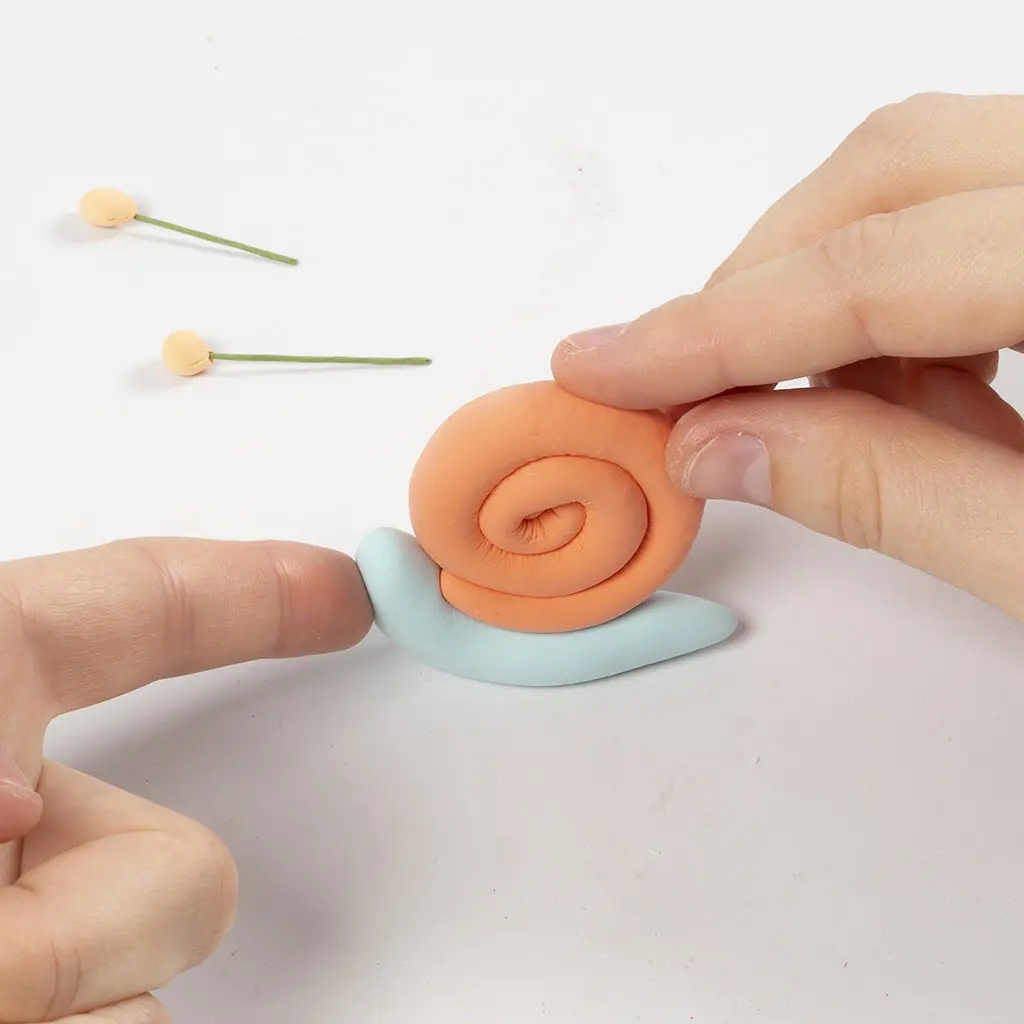 Een vinger drukt het oranje spiraalvormige slakkenhuis vast op het lichtblauwe kleilijfje van de slak.