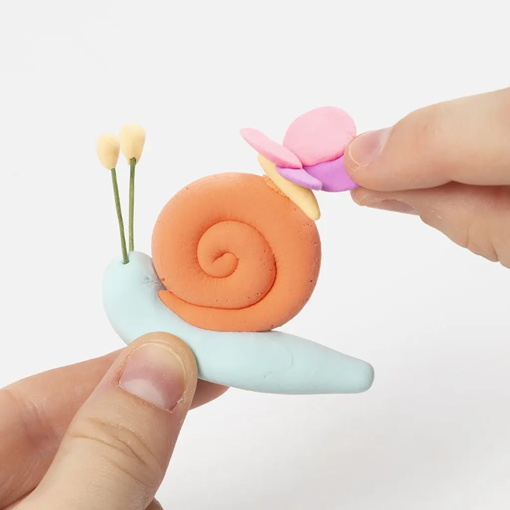 Een hand plaatst een roze-paarse kleivlinder op het oranje slakkenhuis van een lichtblauwe kleislak.