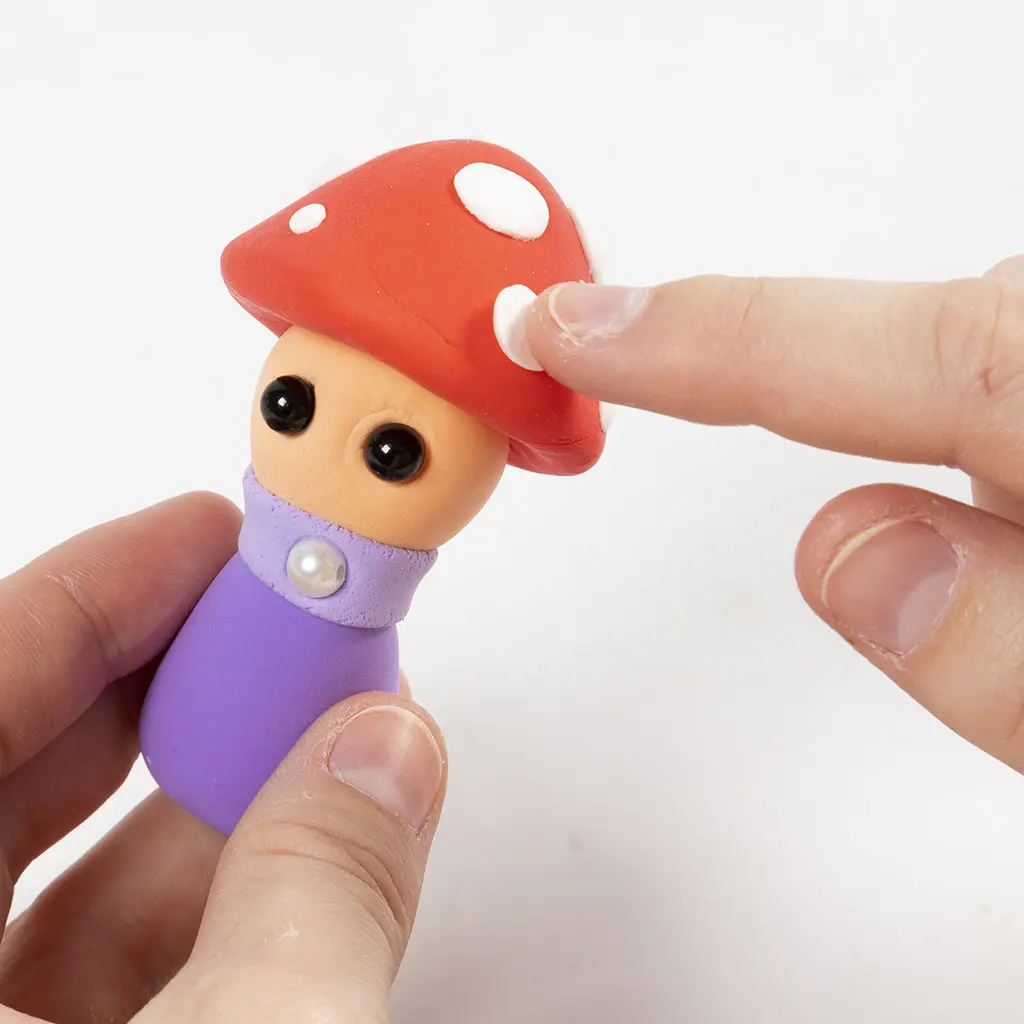 Een hand bevestigt kleine witte kleistippen op het rode hoedje van het in elkaar gezette paddenstoelfiguurtje.